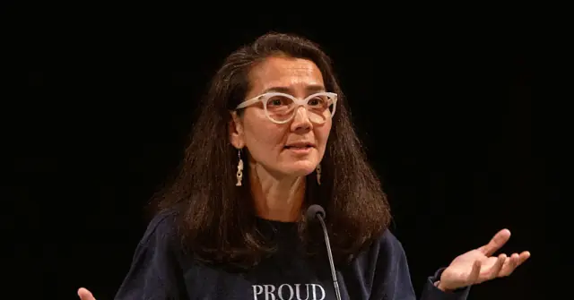 The image shows a person speaking at a podium, wearing a dark shirt with the word "PROUD" visible, and gesturing with their hands.