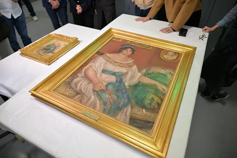 A group of people gather around a large framed painting displayed on a table, with smaller artworks also visible.