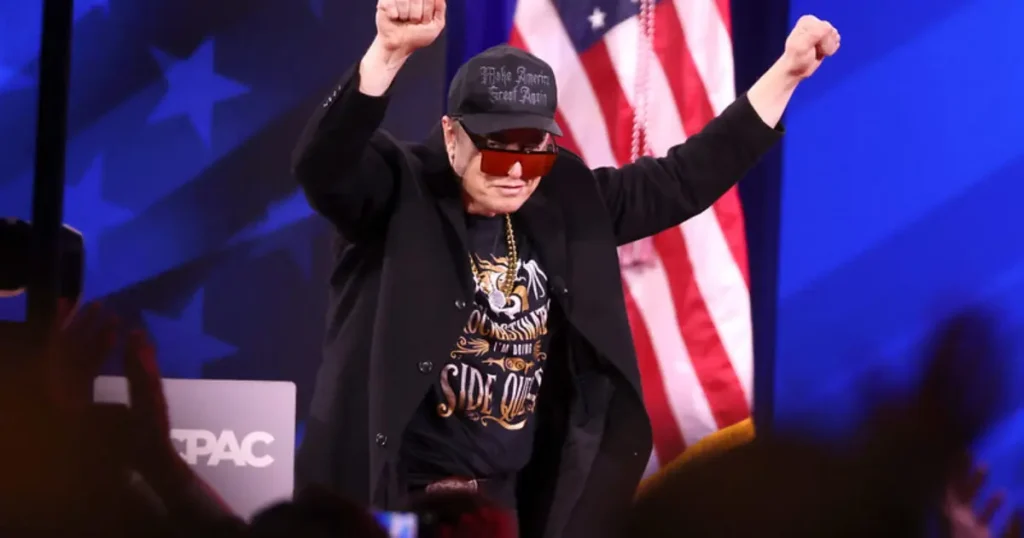A person wearing a "Make America Great Again" hat and sunglasses raises their fists in the air, standing in front of an American flag and a crowd.