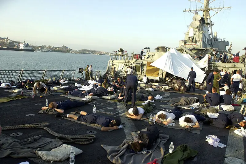 A group of sailors rests on blankets on the deck of a naval ship, with a large ship in the background and a cityscape visible in the distance.