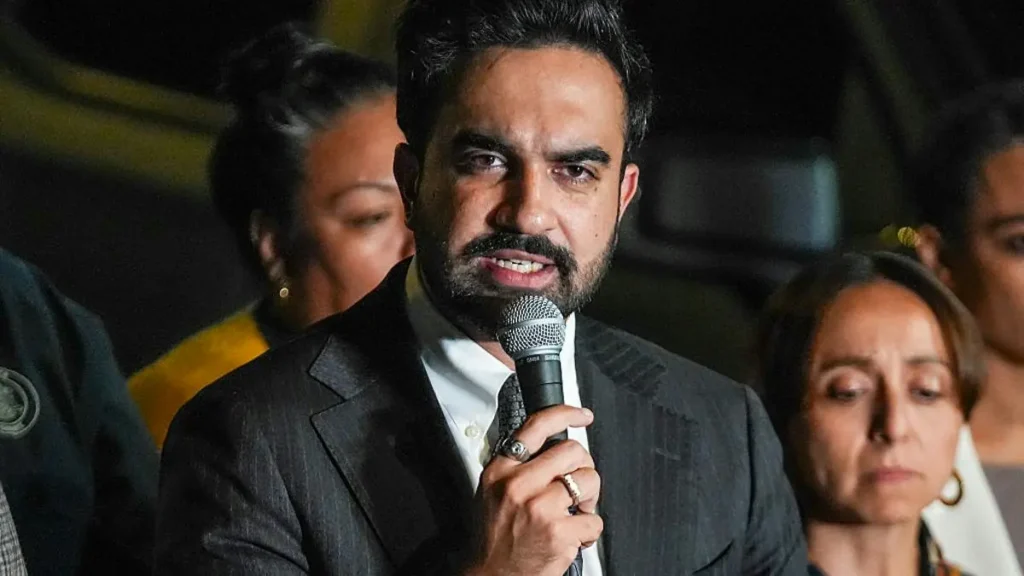 A man in a suit speaks into a microphone at a public event, with an attentive audience in the background.