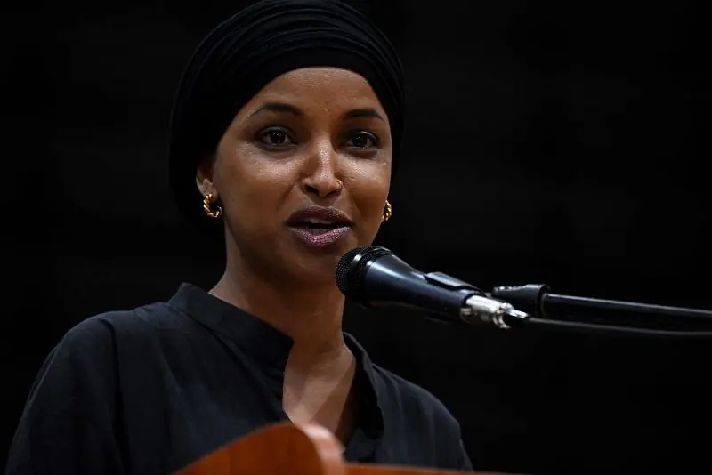 A woman wearing a black headscarf and earrings speaks into a microphone during a public event.