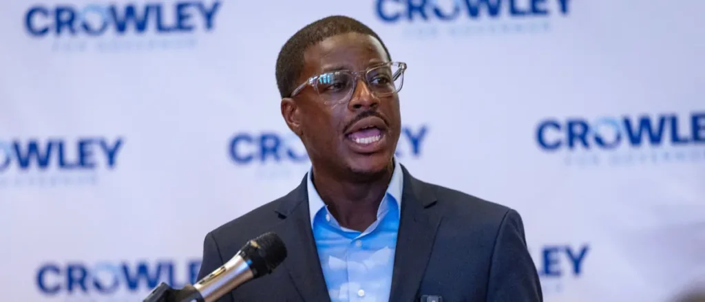 A man in a suit and glasses is speaking into a microphone at a press event with a backdrop featuring the "Crowley" logo.