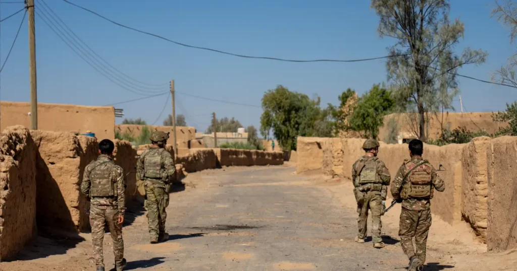 A group of soldiers in camouflage uniforms is seen walking through a narrow, sunlit street lined with mud-brick buildings in a rural area.