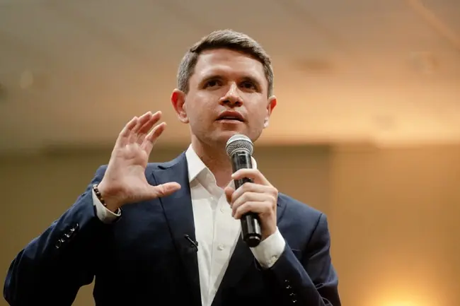 A man in a suit is speaking into a microphone while gesturing with his other hand.