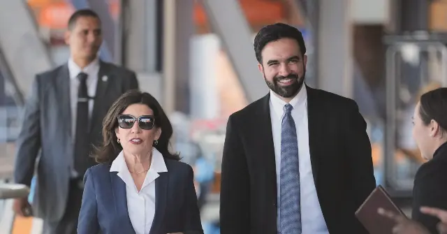 A man and a woman in business attire are walking together, smiling, with a security guard and another person in the background.