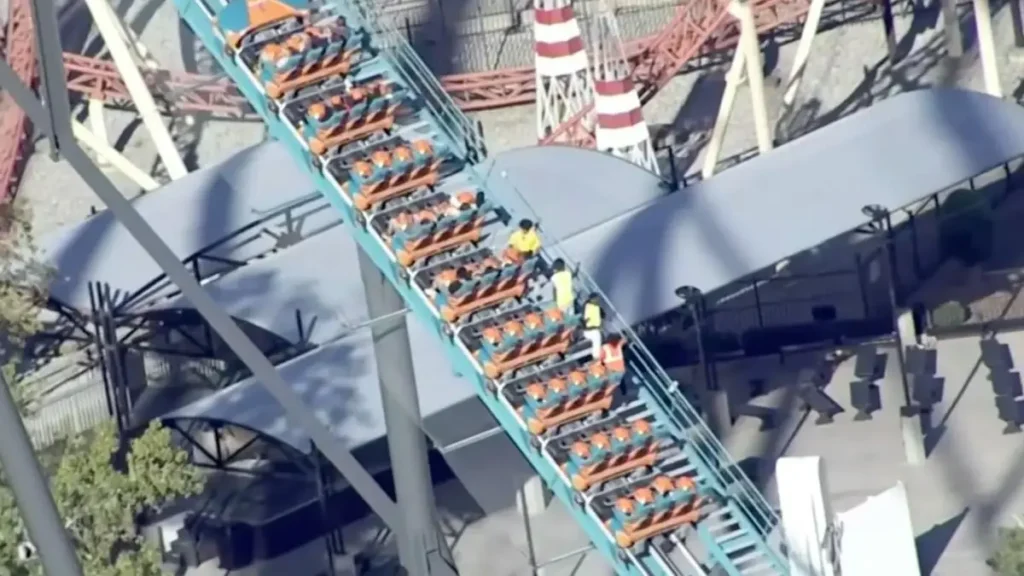 A roller coaster is shown with its cars empty, and a few workers in safety vests are visible on the track.
