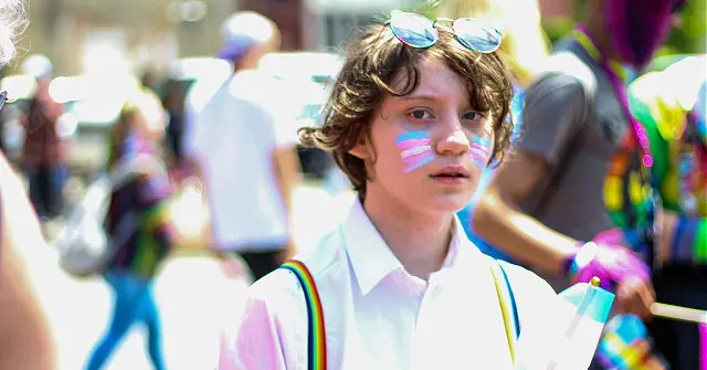 A young person with rainbow-colored face paint and a white shirt stands in a vibrant, colorful crowd, possibly at a pride parade.