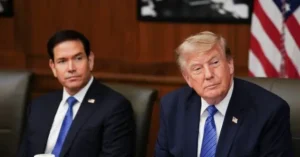 The image shows two men in suits seated in front of an American flag, likely in a formal or official setting.