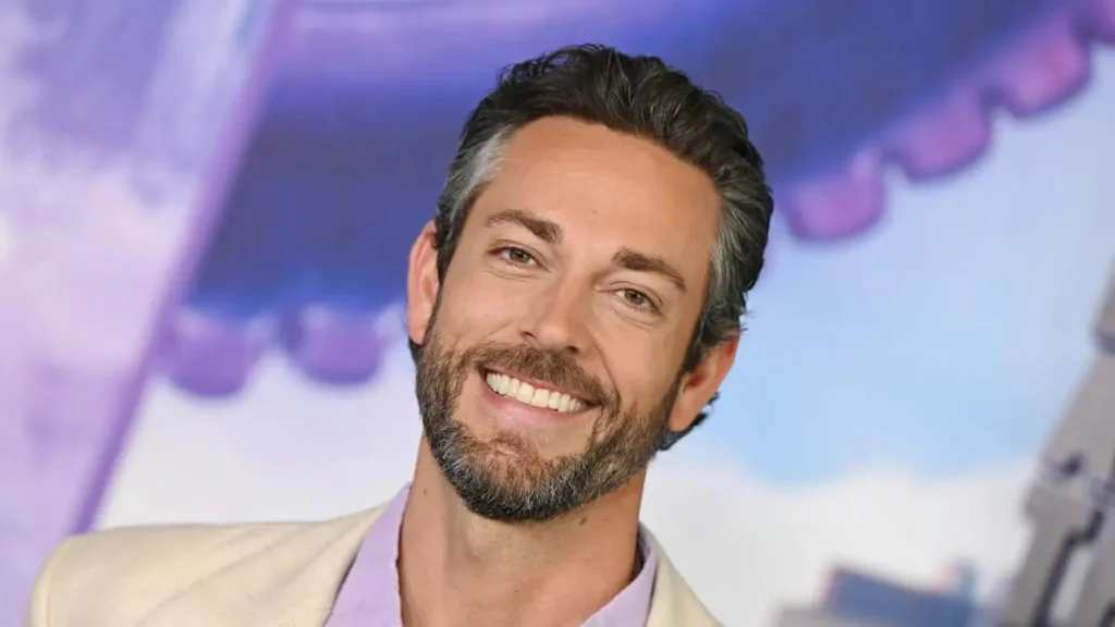 The image shows a man with a beard and short hair, smiling, wearing a light-colored blazer over a purple shirt, with a colorful background.