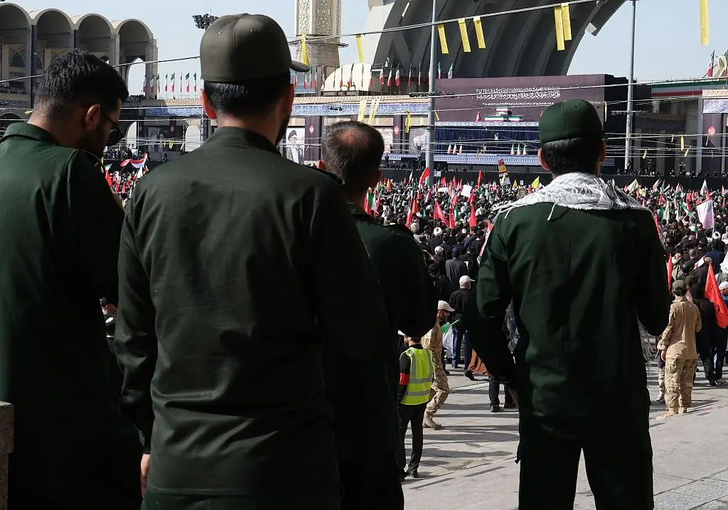 The photo depicts a large gathering of people, many wearing green uniforms, in what appears to be a public square or mosque, with flags and banners visible in the background.