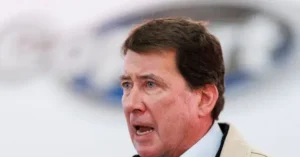 The image shows a man speaking in front of a blurred background featuring a logo.