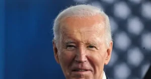 The image shows a close-up of a person with white hair, wearing a suit, against a blue background with blurred circular patterns.