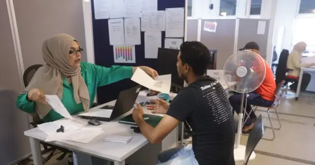 A woman in a hijab and a man are engaged in a discussion at a desk in an office setting, with other individuals working in the background.