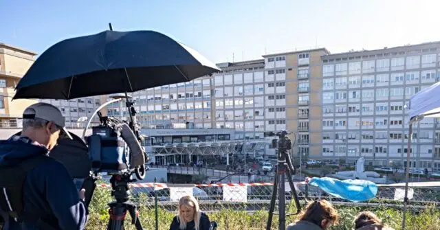A news crew is filming in front of a large building with an umbrella providing shade for the camera equipment.