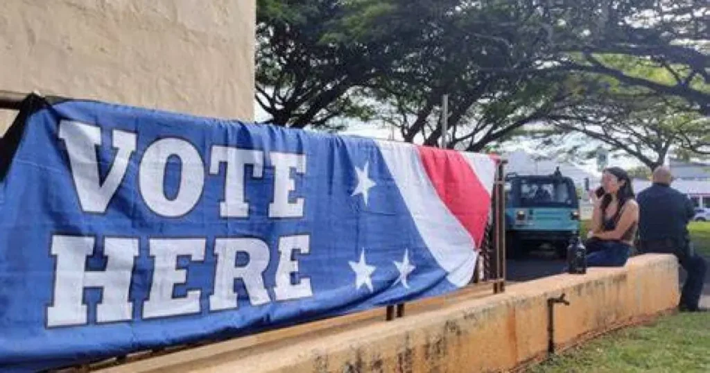 A "Vote Here" sign with a patriotic design is displayed outside a polling station, with a few people sitting nearby.