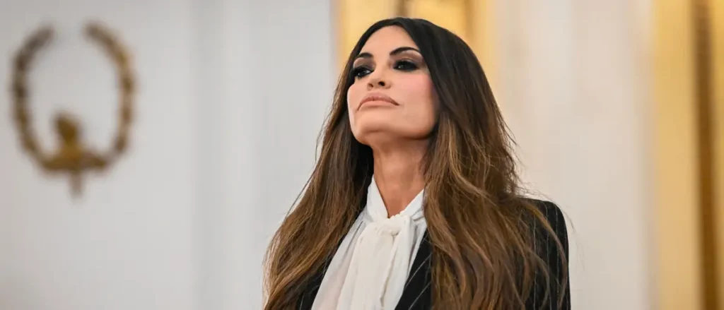 The image shows a person with long, wavy hair wearing a black suit and white blouse, standing in a formal setting with a blurred emblem in the background.