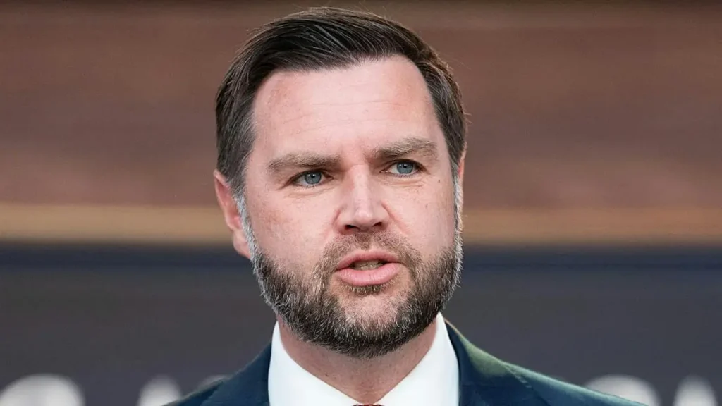 The image shows a man in a suit and tie, speaking or addressing an audience.