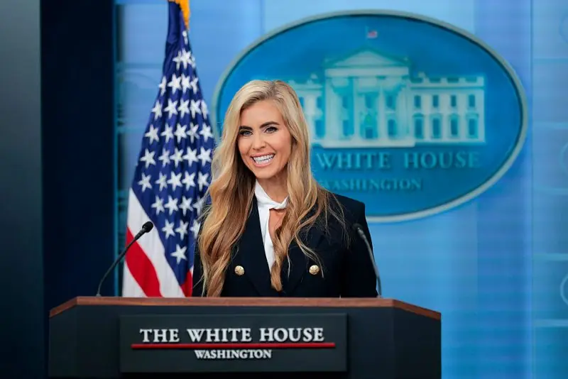 A woman stands at a podium labeled "The White House, Washington" with an American flag and the White House seal in the background.
