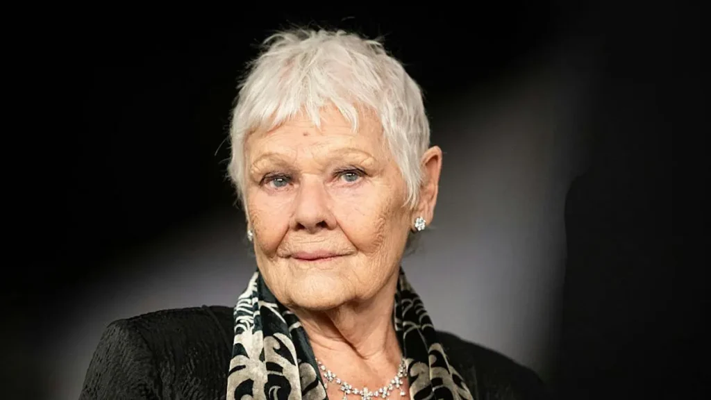 The image shows a person with short, white hair, wearing a black outfit with a patterned scarf and a necklace, set against a dark background.