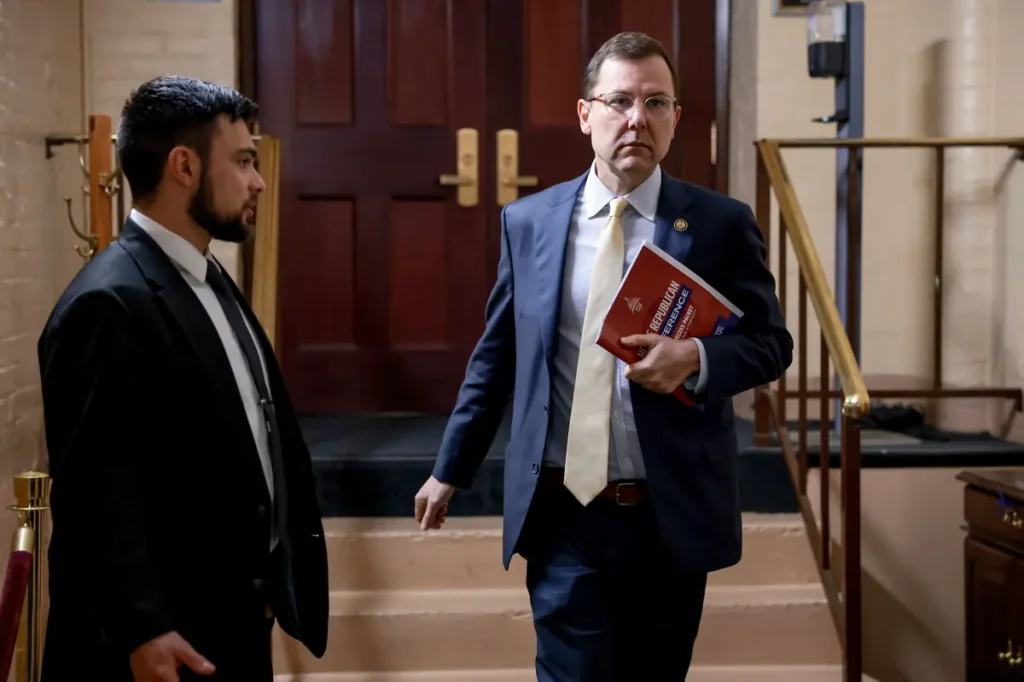 Two men in suits are engaged in a conversation in a formal setting, with one holding a document.