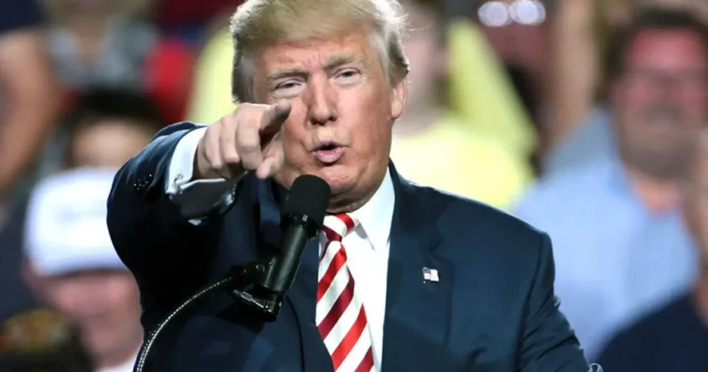 The image shows a person in a suit and tie speaking into a microphone, pointing towards the audience.