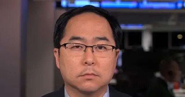 The image shows a man wearing glasses and a suit, likely participating in a news broadcast.