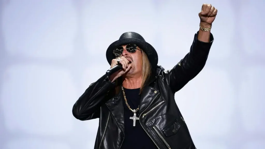 The image shows a performer on stage, wearing a black leather jacket, a cross necklace, and a black hat, holding a microphone and raising one fist in the air.