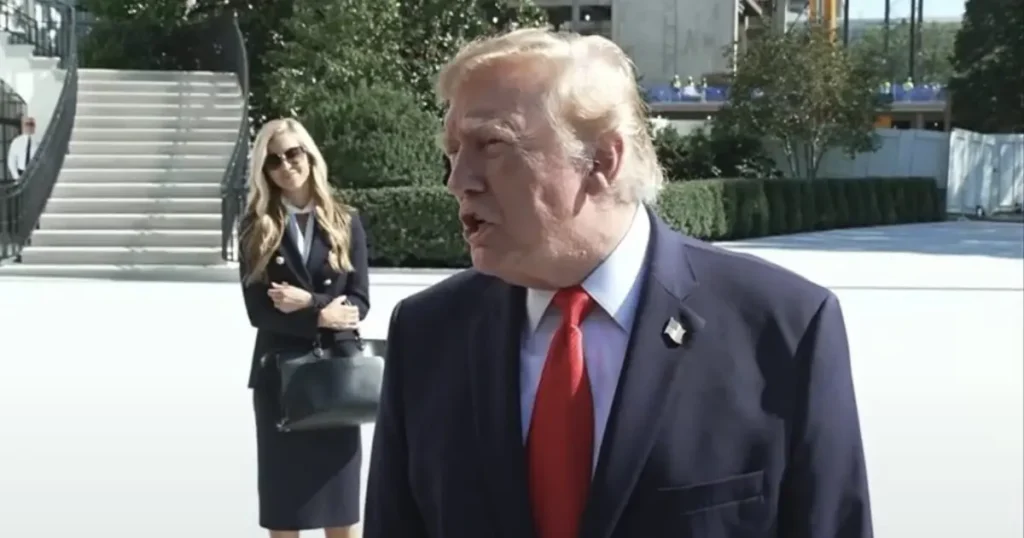 The image shows a man in a suit and red tie speaking outdoors, with a woman in a business suit standing behind him.