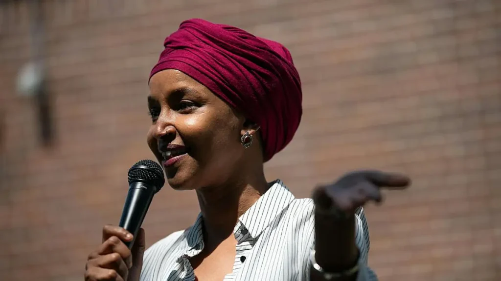 A woman wearing a red turban speaks into a microphone while gesturing with her hand.