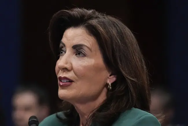 The image shows a woman speaking at a formal event, possibly a hearing or a press conference, as indicated by the microphone and the setting.