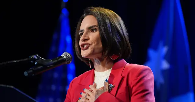 A woman in a red blazer is speaking at a podium with a microphone, with flags in the background.