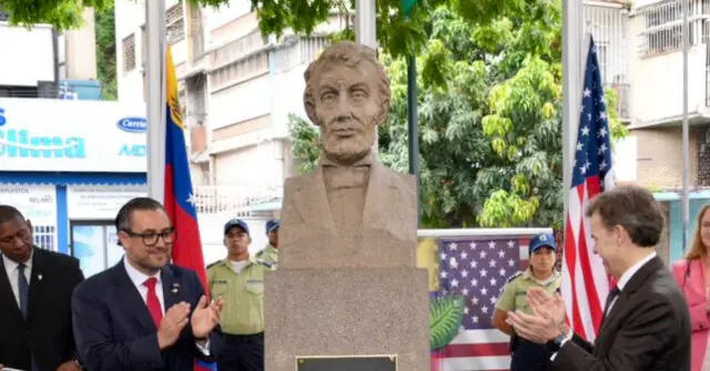 A group of people applaud in front of a bust during a formal event, with flags of Ecuador and the United States in the background.