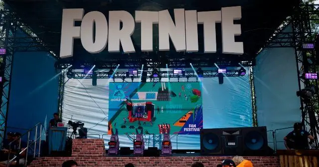 The image shows a stage setup for a Fortnite event, featuring a large screen displaying a map and various characters, with a "FAN FESTIVAL" banner and a stage setup for a presentation or performance.