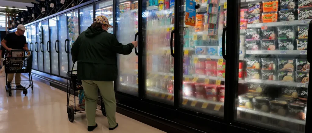 A person is shopping in a grocery store, pushing a cart and looking at the frozen food section.