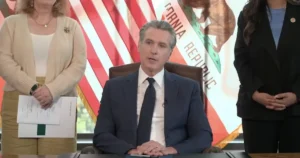 A man in a suit sits at a desk with two women standing behind him, with the California state flag in the background.