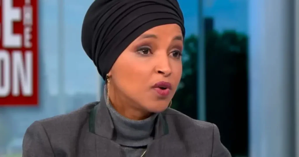 The image shows a person wearing a black headscarf and a gray outfit, speaking on a news program.