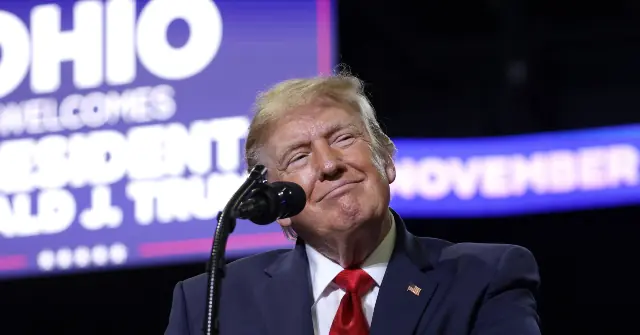 The image shows a person in a suit and tie speaking at a podium with a backdrop that reads "OHIO WELCOMES PRESIDENT DONALD TRUMP NOVEMBER."