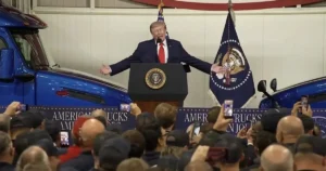 The image shows a political figure speaking at a podium with an American flag and presidential seal in the background, addressing a crowd of people.