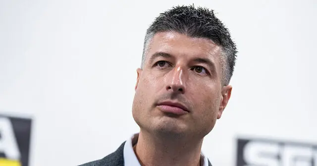 The image shows a man in a suit, looking off to the side with a serious expression, set against a backdrop with logos.