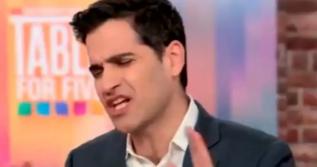 The image shows a man in a suit speaking on a television show set with a colorful background.