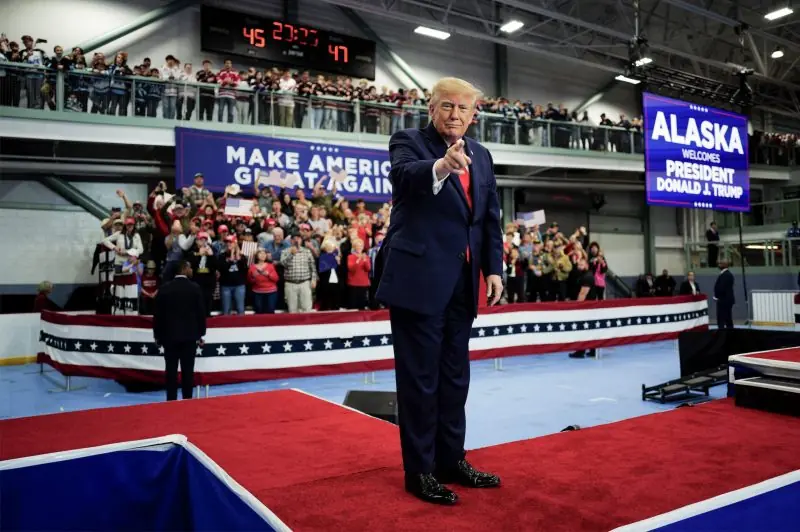 The image shows President Donald J. Trump at a rally in Alaska, where he is pointing towards the crowd, with a sign in the background reading "MAKE AMERICA GREAT AGAIN."