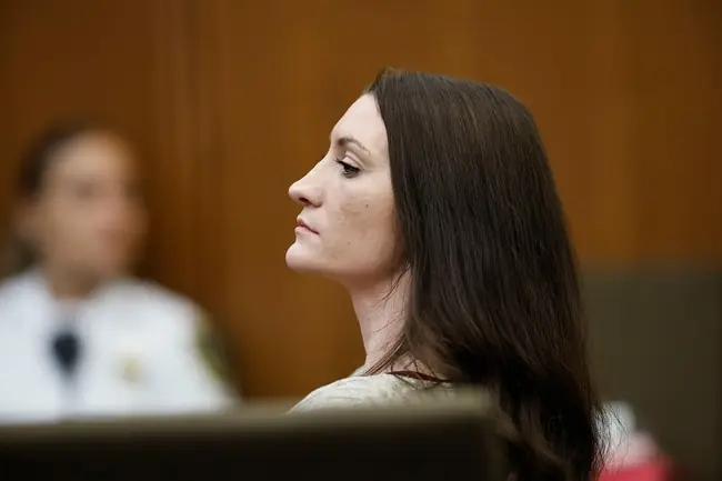 The image shows a woman with long dark hair seated in what appears to be a courtroom setting.