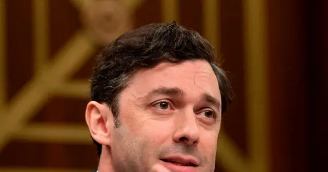 The image shows a close-up of a man with short dark hair, wearing a suit, in what appears to be a formal setting.