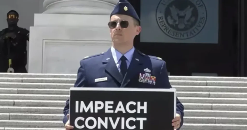 A person in military uniform holds a sign reading "IMPEACH CONVICT" in front of the U.S. House of Representatives building.