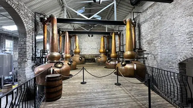 The image shows a distillery with large copper stills and wooden barrels, enclosed by a railing, and set against a backdrop of brick walls and industrial-style lighting.