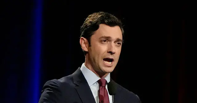 The image shows a man in a suit and tie speaking into a microphone, likely during a formal or professional event.