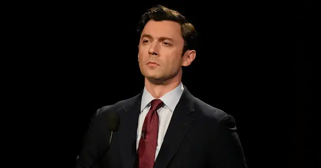 The image shows a man in a suit and tie speaking into a microphone against a dark background.