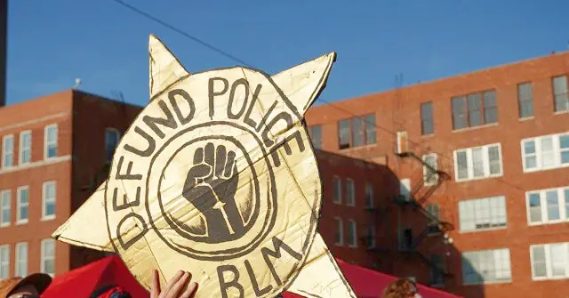 The image shows a protest sign advocating for the defunding of police, with the acronym "BLM" visible, symbolizing the Black Lives Matter movement.