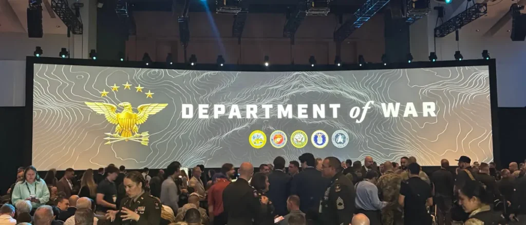 The image shows a large gathering at an event hosted by the Department of War, featuring a prominent display of the U.S. military emblem and various military insignias.